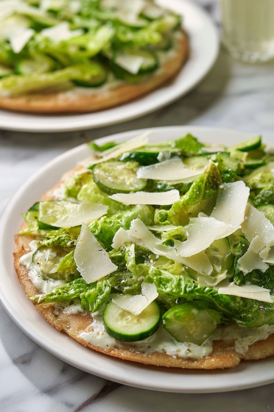 Viral Chicken Crust Caesar Salad Pizza, low-carb chicken crust pizza, healthy Caesar salad pizza, easy chicken crust pizza recipe, gluten-free chicken pizza - The dish shows a flat round base with a light brown, slightly crispy texture. On top of the base, there is a layer of creamy white sauce spreading evenly. Above the sauce, there is a thick layer of green romaine lettuce pieces, mixed with round slices of light green cucumber scattered on top. Thin, irregular white shavings, likely cheese, are spread across the green vegetables. The dish is served on a simple white plate placed on a white marbled surface, with another similar dish visible blurred in the background. photo taken with an iphone --ar 2:3 --v 7