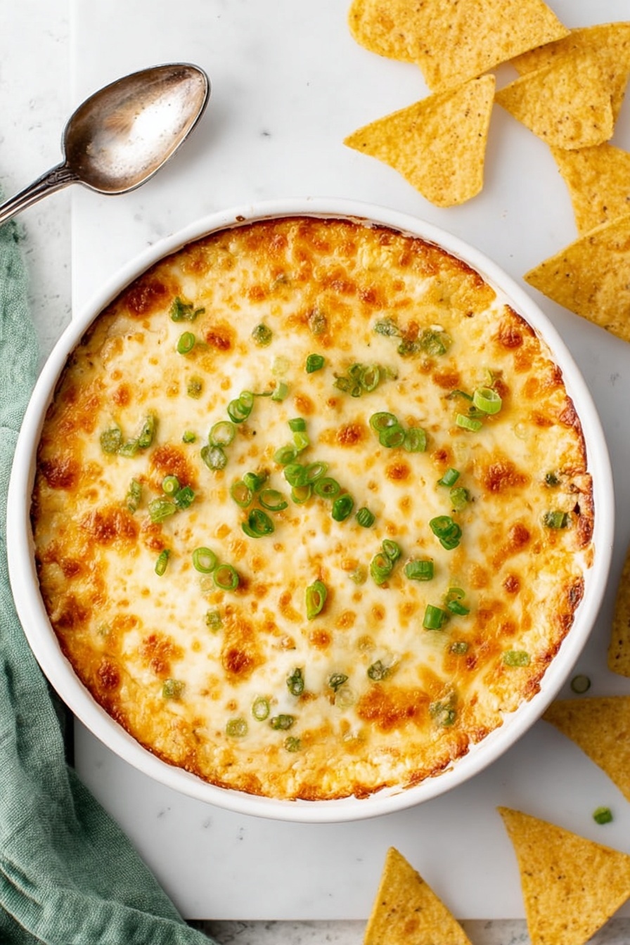 Crab Rangoon Dip with Crispy Wonton Chips, Crab Rangoon Dip, Wonton Chip Appetizer, Easy Crab Dip Recipe, Party Crab Dip - A round white baking dish filled with a golden, bubbly cheese layer on top, showing patches of light brown where the cheese is slightly toasted, scattered with small rings of bright green chopped scallions. The entire dish sits on a white marbled surface with a vintage silver spoon to the side, broken and whole triangular yellow tortilla chips nearby, and a soft green cloth partly visible at the edge. Photo taken with an iphone --ar 2:3 --v 7