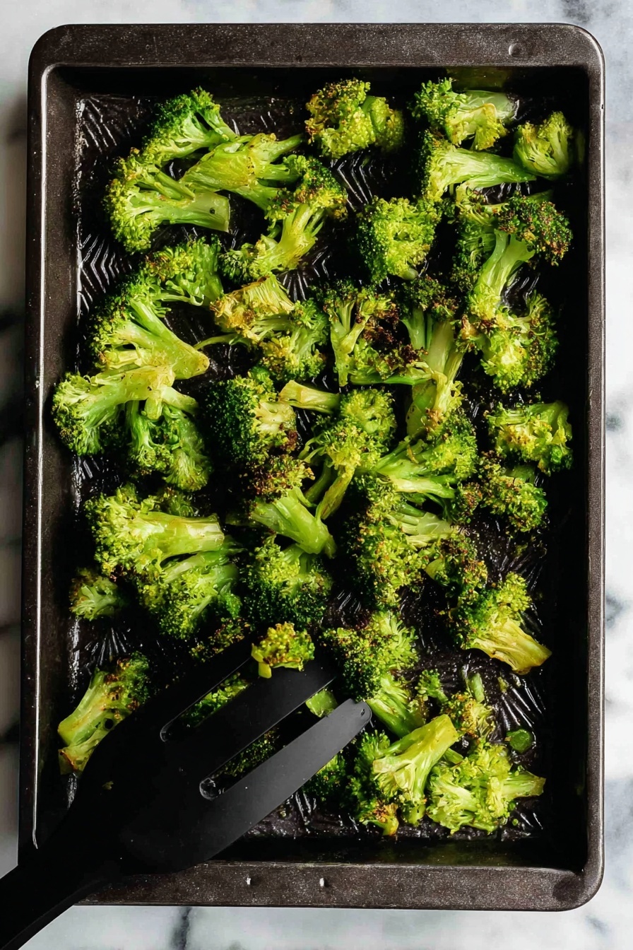 Oven Roasted Broccoli with Spices, roasted broccoli side dish, spicy broccoli recipe, garlic paprika broccoli, healthy roasted vegetable - A dark rectangular baking tray holds a single layer of cooked broccoli florets. The broccoli pieces are bright green with some slightly browned tips, showing a crispy texture. The florets vary in size and shape, spread unevenly across the tray. In the bottom right corner, a black spatula is lifting some broccoli. The tray rests on a white marbled surface. photo taken with an iphone --ar 2:3 --v 7
