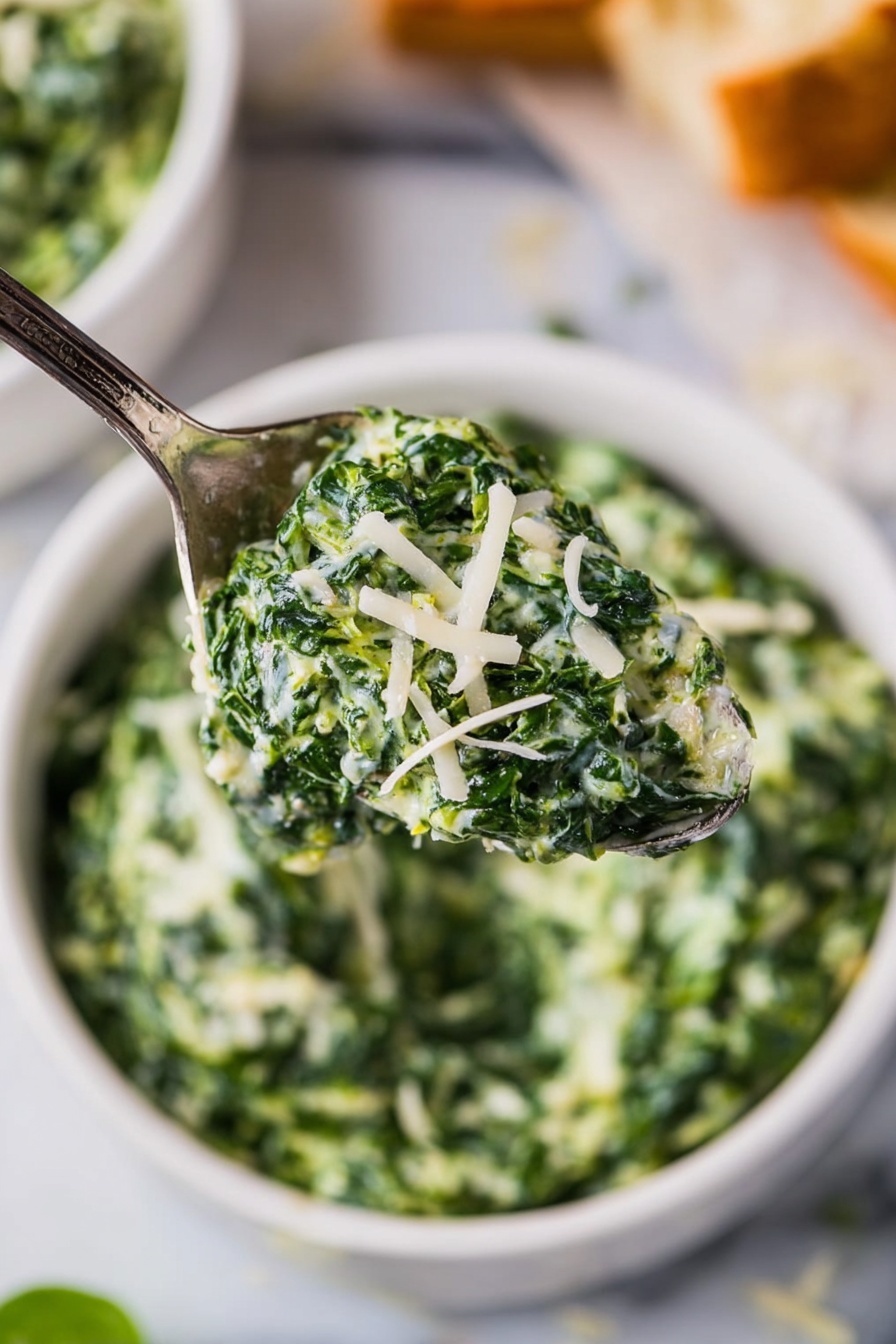 Creamed Spinach, creamed spinach side dish, cheesy creamed spinach, easy creamed spinach recipe, creamy spinach sides - The image shows a spoon holding a creamy green spinach mixture with small curls of white cheese sprinkled on top. Below the spoon, there is a white bowl filled with the same creamy spinach, and the bowl sits on a white marbled surface. Around the bowl, there are blurred out pieces of bread in the background, adding a warm brown color contrast to the green and white of the spinach dish. The textures of the spinach are soft and creamy with bits of shredded cheese visible. Photo taken with an iphone --ar 2:3 --v 7