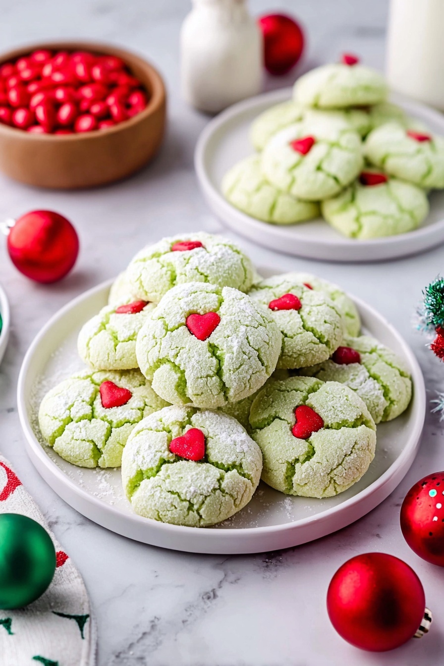 Green Peppermint Cake Mix Cookies, peppermint cookie recipe, holiday cake mix cookies, easy peppermint cookies, festive minty cookies - A large pile of light green cookies with a soft cracked texture, each topped with a small red heart candy, sits on a white plate in the center. Two smaller white plates in the background hold more of the same cookies. The cookies are dusted lightly with powdered sugar. Around the plates are red and green round Christmas ornaments and a small wooden bowl filled with red candies on a white marbled surface. A white bottle is visible in the background. Photo taken with an iphone --ar 2:3 --v 7