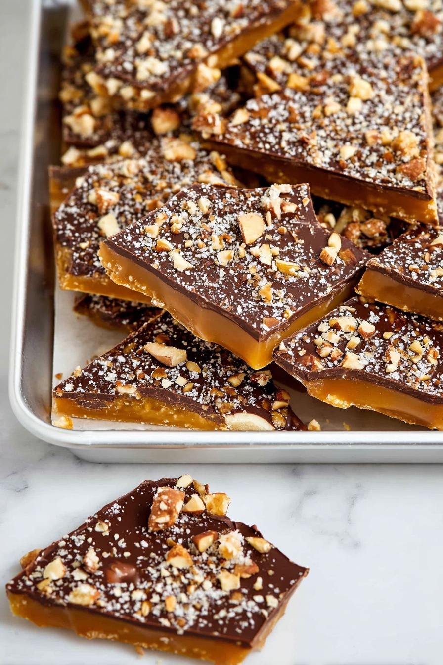 Homemade Toffee with Chocolate and Pecans, Toffee recipe with chocolate and pecans, easy toffee with nuts and chocolate, homemade brittle with pecans, caramel toffee dessert - The image shows a white tray full of irregular triangular pieces of candy with three visible layers: a bottom layer of golden caramel, a shiny middle layer of smooth dark chocolate, and a top layer sprinkled heavily with crushed nuts in light brown and white tones. Next to the tray are two more pieces of the same candy, one fully visible and one partly shown, lying on a white marbled surface. The overall look is rich and textured with the nut pieces creating a rough surface on top of the smooth chocolate layer photo taken with an iphone --ar 2:3 --v 7