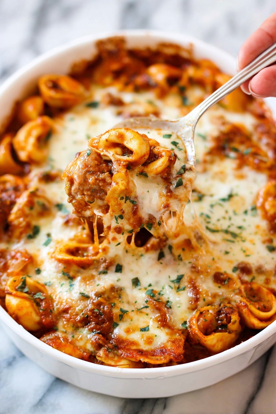 Cheesy Baked Tortellini with Beef, baked tortellini with beef, cheesy stuffed pasta bake, easy cheesy beef casserole, comfort food dinner recipes - A white dish filled with baked tortellini pasta tossed in a rich red tomato sauce and covered with a thick layer of melted, golden-brown cheese with green herb sprinkles on top. Some tortellini are visible under the cheese, showing their smooth curved shapes and orange sauce coating. A woman's hand holds a silver spoon lifting a portion of the cheesy pasta, with melted cheese stretching slightly. The background shows more pasta inside the white dish on a white marbled surface. photo taken with an iphone --ar 2:3 --v 7