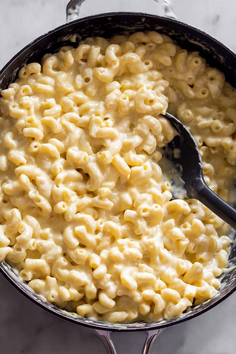 Creamy Baked Mac and Cheese, baked mac and cheese recipe, cheesy baked pasta, homemade mac and cheese, comfort food recipes - The image shows a black pan filled with creamy macaroni and cheese. There is one thick layer of soft elbow pasta coated in smooth, light yellow cheese sauce. A black spoon is partly submerged in the macaroni, lifting a scoop on one side of the pan. The pan is set on a white marbled surface which is slightly visible around the edges. Photo taken with an iphone --ar 2:3 --v 7