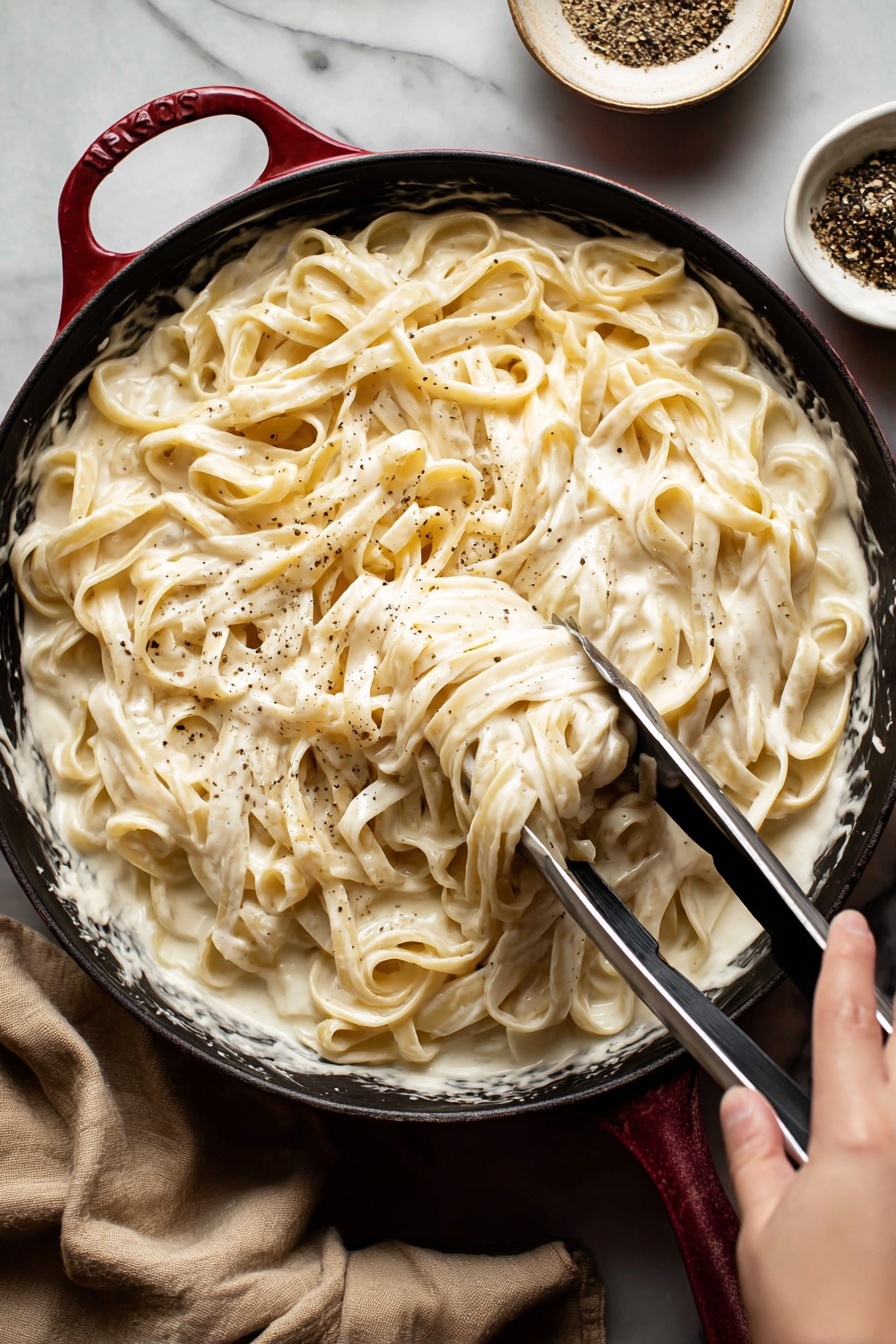 Vegan Cashew Alfredo Sauce, dairy-free Alfredo sauce, plant-based Alfredo recipe, vegan pasta sauce, dairy-free creamy sauce - A close-up view of creamy fettuccine pasta served in a black pan with a red handle edge, the pasta is covered in a thick, smooth white sauce that looks rich and velvety; the long pasta strands are mixed well with the sauce and lightly sprinkled with black pepper; a woman's hand is holding metal tongs, lifting some pasta from the pan; in the background, there is a small white bowl with black pepper, and the whole setting is on a white marbled surface with a beige cloth nearby photo taken with an iphone --ar 2:3 --v 7