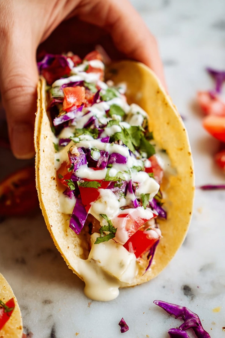 Vegan Fish Tacos with Fresh Toppings, vegan fish tacos, plant-based fish tacos, vegan taco recipes, healthy vegan tacos - A small soft taco held by a woman's hand shows three clear layers: the bottom layer is a pale yellow soft tortilla, the middle layer consists of white cheese chunks and small pieces of red tomato, and the top layer has deep purple cabbage pieces, small green bits of cilantro, and a drizzle of creamy white sauce covering the ingredients. The taco is placed on a white marbled surface with some small scattered pieces of purple cabbage and red tomato around it. Photo taken with an iphone --ar 2:3 --v 7