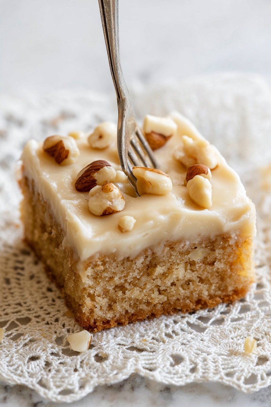 Peanut Butter Cake with Creamy Frosting, peanut butter cake, moist peanut butter cake, creamy frosting recipe, homemade peanut butter dessert - A close-up of a single square piece of cake with two layers, the bottom layer is a light brown, soft, crumbly cake base, and the top layer is a thick, pale cream frosting with a smooth texture. The frosting is decorated with whole, shiny brown and white nuts scattered unevenly on top. A fork is pressing slightly into the right side of the frosting. The cake rests on a delicate crochet lace doily with intricate patterns, placed on a white marbled surface. Photo taken with an iphone --ar 2:3 --v 7