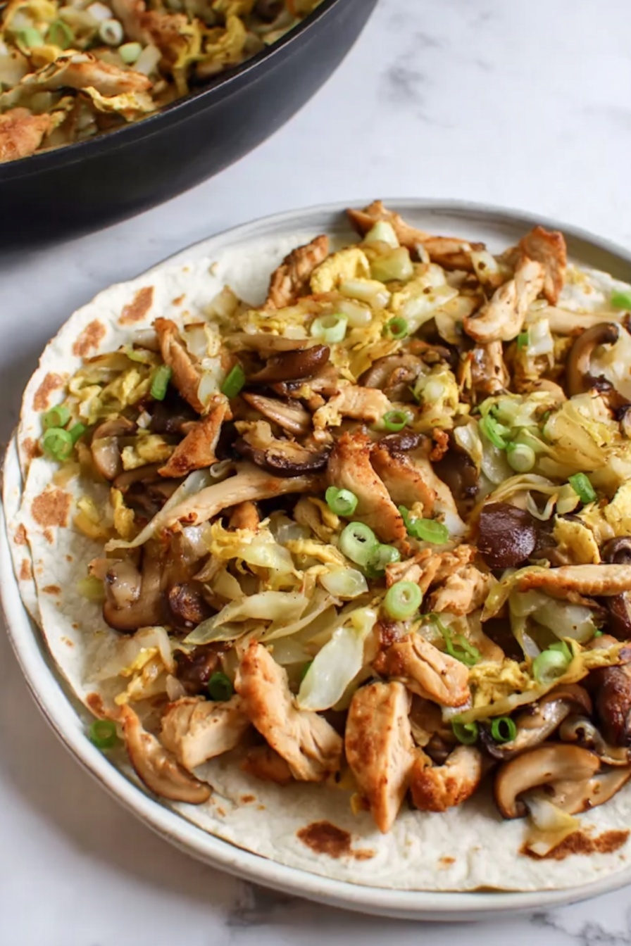 Moo Shu Chicken Stir-Fry, Chinese chicken stir-fry, quick stir-fry recipes, healthy chicken dinner, homemade stir-fry meal - This image shows a close-up of a black frying pan filled with a stir-fry dish. The dish has many pieces of brown, cooked chicken strips mixed with light yellow cabbage slices and orange-yellow egg strips. There are also some sliced mushrooms with a dark brown outer edge and light beige inside, scattered throughout. Small green onion pieces are sprinkled over the dish adding a splash of green color. The frying pan is set on a white marbled surface. photo taken with an iphone --ar 2:3 --v 7