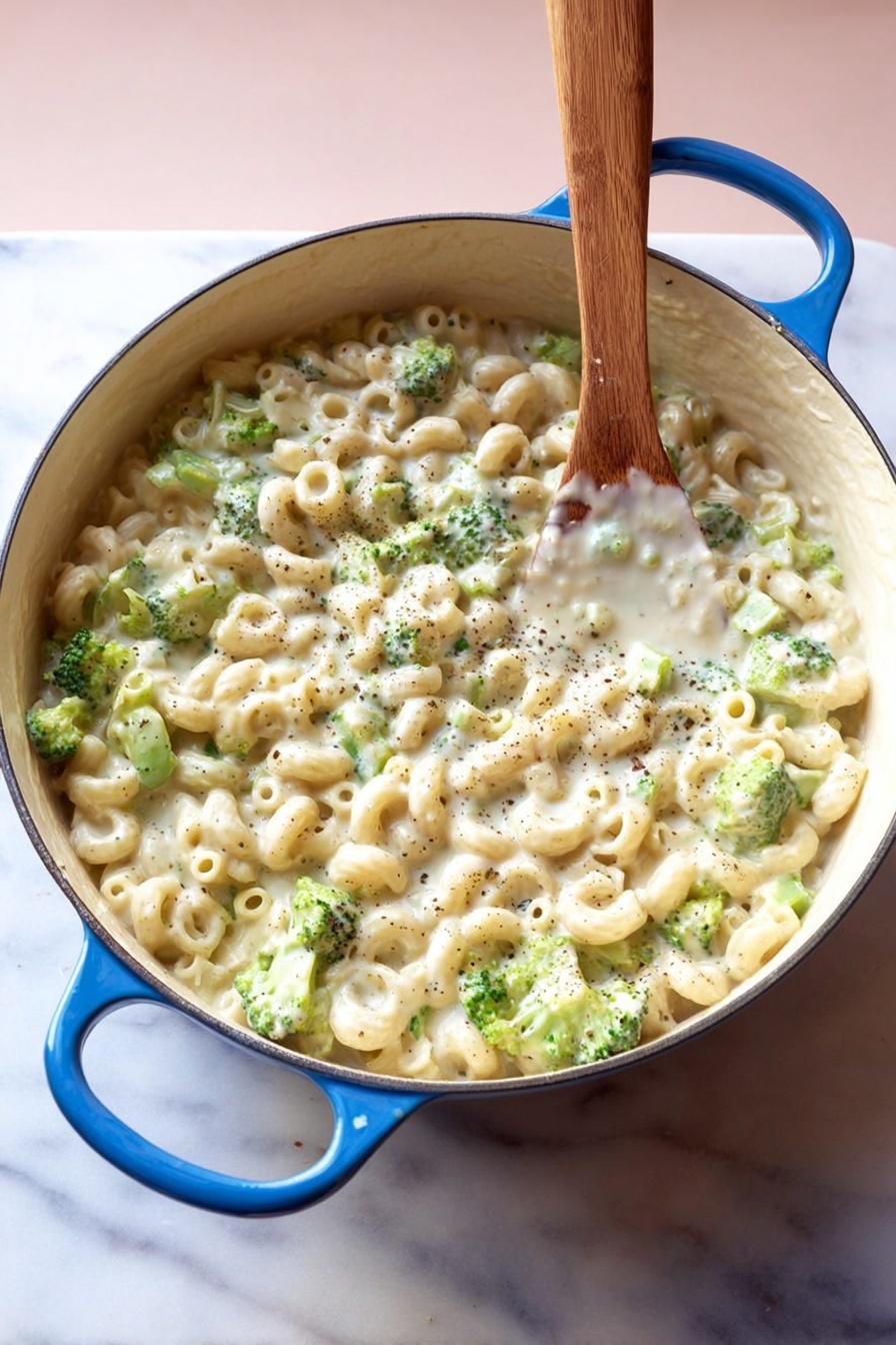Creamy Broccoli Pasta, creamy broccoli pasta recipe, cheesy broccoli pasta, easy broccoli pasta dinner, quick vegetarian pasta - A large white pot with blue handles is filled with small tube pasta and small green broccoli pieces mixed in a thick, creamy white sauce with visible black pepper specks, all stirred by a wooden spoon standing upright in the pot, placed on a white marbled surface with a soft pink background, photo taken with an iphone --ar 2:3 --v 7