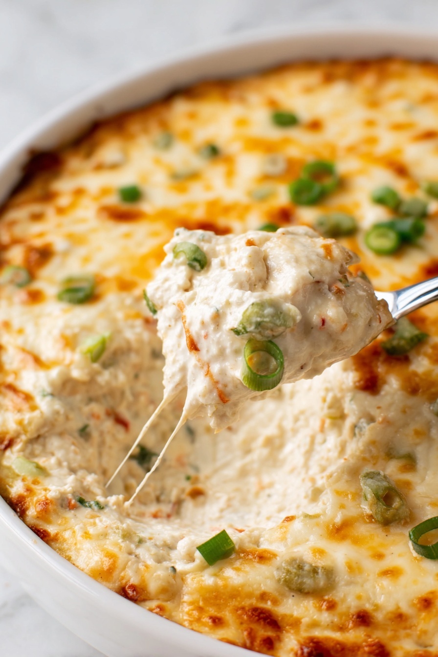 Crab Rangoon Dip with Crispy Wonton Chips, Crab Rangoon Dip, Wonton Chip Appetizer, Easy Crab Dip Recipe, Party Crab Dip - A close-up of a white dish filled with a creamy dip that has a thick melted cheese layer on top. The cheese is golden with light brown spots and sprinkled with small green onion pieces. The creamy layer underneath is light beige with visible small bits of other ingredients, showing a rich and creamy texture. A spoon is lifting a scoop of the dip, revealing the thick inside. The dish is on a white marbled surface. photo taken with an iphone --ar 2:3 --v 7