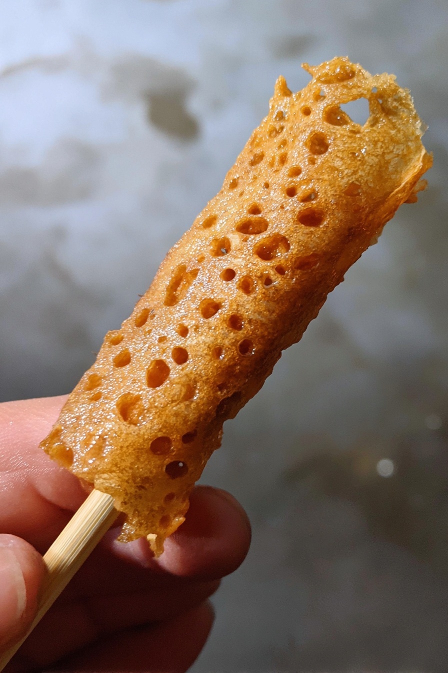 Brandy Snaps, easy Brandy Snaps, homemade Brandy Snaps, holiday Brandy Snaps, crispy Brandy Snaps - A close-up image of a golden brown, thin, lace-like fried snack wrapped around a light wooden stick held by a woman's hand, with a textured, crispy surface that has small holes and a shiny finish, against a background with a white marbled texture. photo taken with an iphone --ar 2:3 --v 7