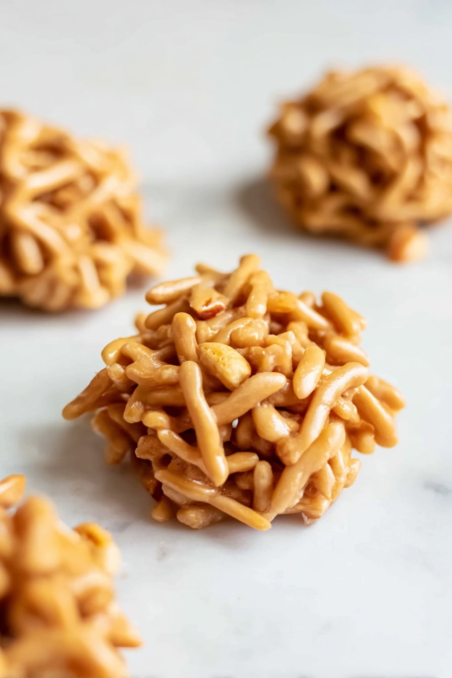 No Bake Haystack Cookies with Peanuts, easy no bake cookies, crunchy peanut cluster recipe, quick no bake snacks, homemade haystack cookies - The image shows small round clusters made of light brown, thin, stick-like pieces intertwined with small peanut bits visible in the mix. Each cluster is a single layered mound, with a rough and irregular texture, placed on a smooth white marbled surface. There are three clusters scattered, with one in clear focus at the front and the other two slightly blurred in the background. The sticks have a matte finish with subtle shadows giving depth and softness to the clusters photo taken with an iphone --ar 2:3 --v 7