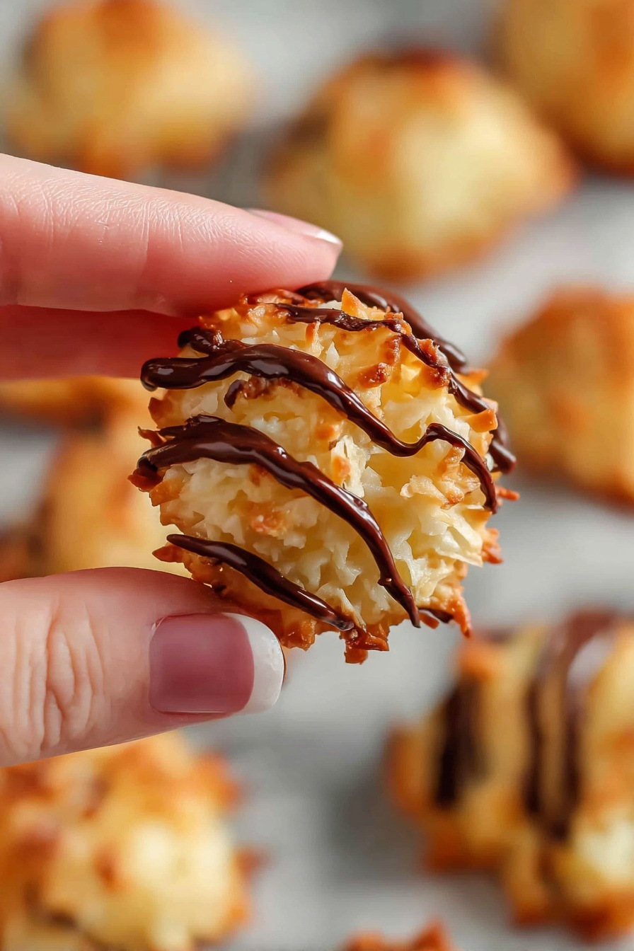 Coconut Macaroons with Condensed Milk, easy coconut macaroons, no-bake coconut treats, chewy coconut cookies, homemade coconut desserts - A close-up of a small round cookie made of shredded coconut with a rough, golden-brown surface, topped with several thick dark chocolate drizzles running diagonally across it; the cookie is held delicately between the thumb and woman’s hand index finger, and in the blurry background, more similar cookies are spread out on a white marbled surface. photo taken with an iphone --ar 2:3 --v 7