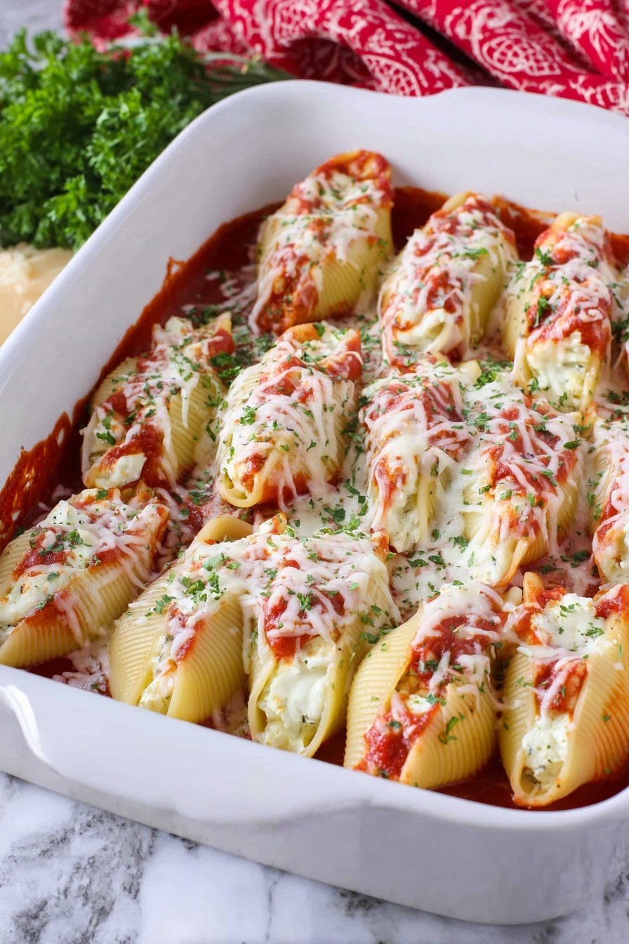 Cheesy Chicken Stuffed Shells, cheesy stuffed pasta, chicken pasta bake, baked stuffed shells, creamy chicken pasta - The image shows a white baking dish filled with two layers of large pasta shells. The bottom layer is covered with a red tomato sauce, and the shells are stuffed with white cheese inside, visible in some openings. The top layer consists of more pasta shells placed closely next to each other, covered with melted white cheese sprinkled with green herbs and bits of red tomato sauce. The dish is placed on a white marbled surface, with some green herbs and a red patterned cloth in the background. Photo taken with an iphone --ar 2:3 --v 7