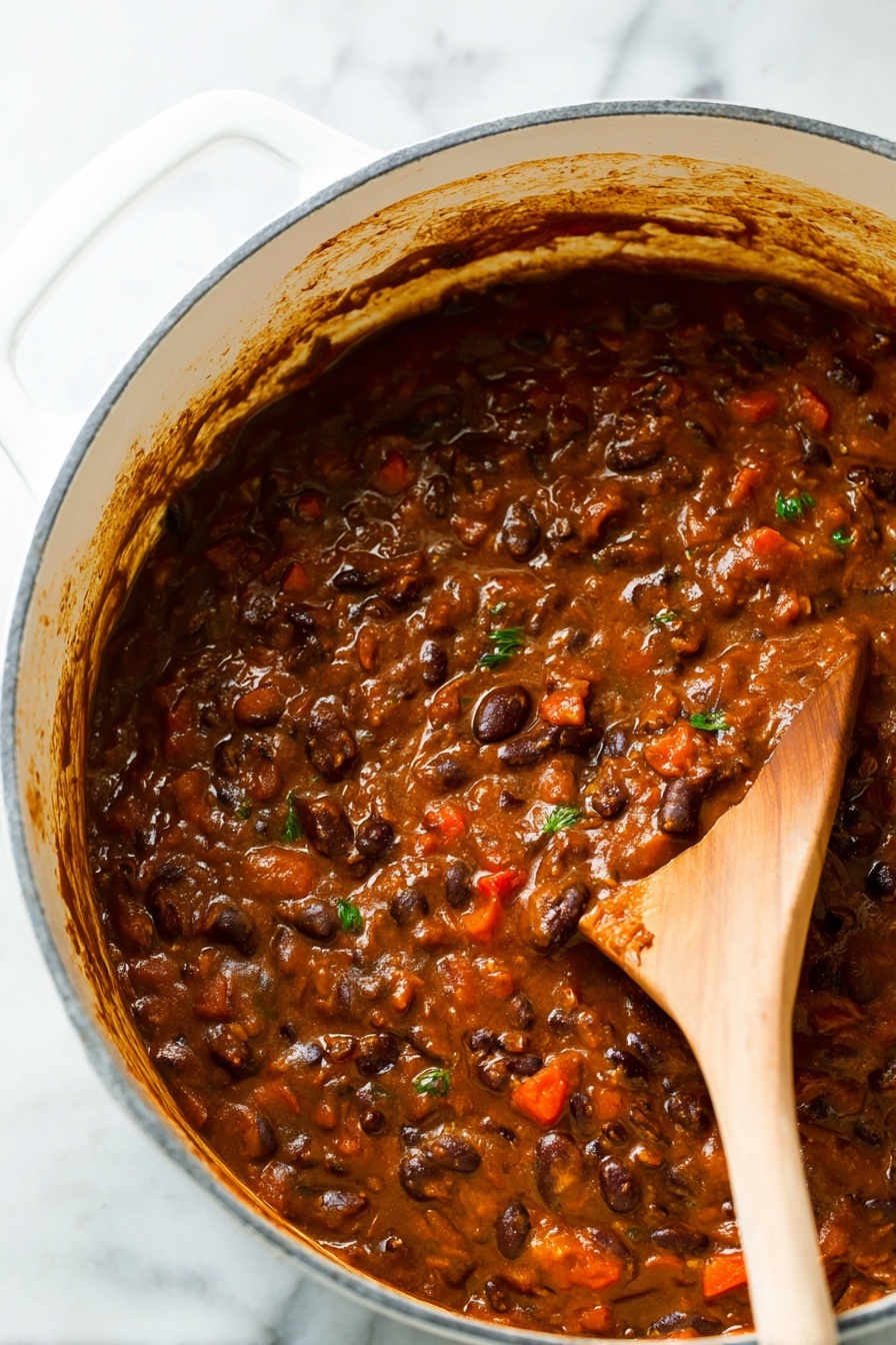 Vegetarian Chili, vegetarian chili recipe, plant-based chili, healthy vegetarian dinner, quick vegetarian chili - A close-up view of a thick stew-like dish inside a large white pot with a white marbled surface underneath. The dish has a rich brown color with visible layers of ingredients, including dark brown beans, small red chunks, and some green herb bits mixed throughout. A wooden spoon is partly dipped in the stew on the right side, showing the texture which is soft and slightly chunky with a glossy finish. The sides of the pot have some sauce splashes and the scene looks warm and hearty. Photo taken with an iphone --ar 2:3 --v 7