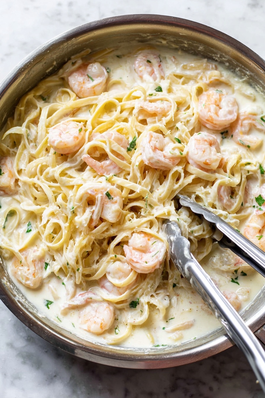 Shrimp Alfredo Pasta, creamy shrimp pasta, easy shrimp Alfredo, quick pasta recipes, shrimp pasta dinner - A close-up view of a silver pan filled with creamy white sauce pasta mixed with pink shrimp. The pasta, light yellow and smooth, is covered in a thick white sauce and evenly spread throughout the pan. Plump shrimp are scattered on top and mixed in, their soft pink color contrasting with the white sauce. Small green herb pieces are sprinkled over the dish for a fresh touch. A pair of metal tongs and a spoon are resting on the pasta, indicating the dish is being stirred or served. The pan sits on a white marbled surface. photo taken with an iphone --ar 2:3 --v 7