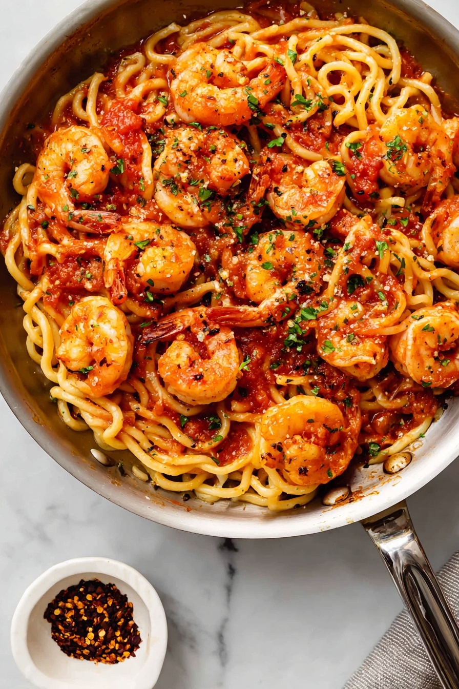 Shrimp Fra Diavolo, spicy seafood pasta, quick seafood dinner, Italian shrimp dish, flavorful shrimp recipes - The image shows a black pan full of curly pasta mixed with cooked shrimp. The pasta is light yellow with a slightly wavy texture, while the shrimp are orange with reddish highlights, laid on top and mixed in all around. There is a red tomato sauce spread unevenly on the pasta and shrimp, and small bits of green herbs sprinkled across the dish. The pan is on a white marbled surface with a small white bowl of red chili flakes beside it at the bottom left. The metal handle of the pan extends out to the right side. Photo taken with an iphone --ar 2:3 --v 7