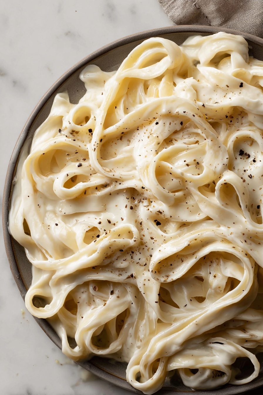 Vegan Cashew Alfredo Sauce, dairy-free Alfredo sauce, plant-based Alfredo recipe, vegan pasta sauce, dairy-free creamy sauce - This image shows a close-up view of wide fettuccine noodles covered in a thick, creamy white sauce. The sauce looks smooth and rich, evenly coating the noodles. Light brown small specks of black pepper are scattered across the pasta, adding texture and contrast. The dish fills the frame, with noodles tangled and folded over each other in layers. The background is a white marbled surface. photo taken with an iphone --ar 2:3 --v 7