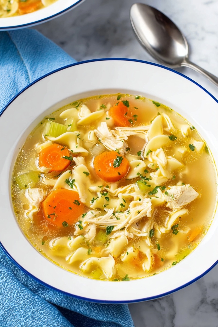 Vegan Chicken Noodle Soup, plant-based chicken noodle soup, vegan comfort food recipes, dairy-free chicken noodle soup, healthy vegan soup recipes - A bowl of chicken noodle soup is shown with clear, yellowish broth filling the white bowl with a thin blue rim. The soup has several layers including bright orange carrot slices and pale green celery pieces scattered throughout. Soft, wavy egg noodles are curled in different shapes, mixed with small chunks of cooked chicken that are light beige with a tender texture. Small green parsley flakes are sprinkled on top, adding a fresh touch. The bowl sits on a white marbled surface with a blue cloth and shiny silver spoon nearby. photo taken with an iphone --ar 2:3 --v 7