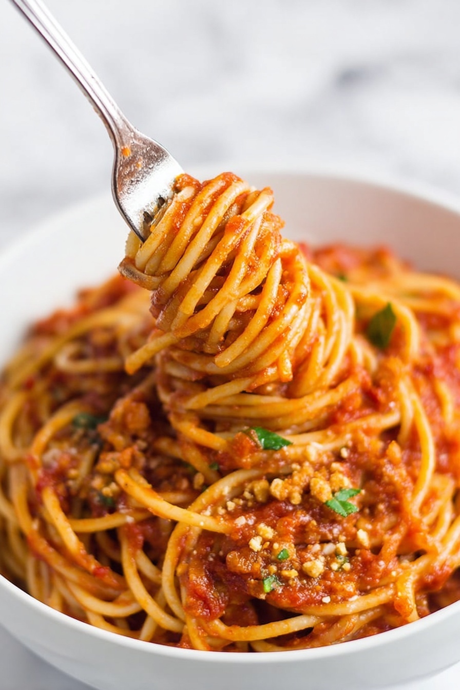 Instant Pot Lentil Spaghetti, Lentil Spaghetti Recipe, Healthy Lentil Pasta, Easy Instant Pot Dinner, Protein-Packed Pasta Dish - A close-up of a white bowl filled with spaghetti noodles coated in rich red tomato sauce with visible small chunks and texture throughout. The spaghetti is thick and twisted around an inserted fork in the center, lifting the noodles upward, showing a dense mix of sauce and soft pasta strands. Small bits of green herbs and crumbled light brown topping are scattered on the surface, adding color contrast. The bowl is placed on a white marbled surface, and the image has a soft background with a focus on the noodles and sauce. photo taken with an iphone --ar 2:3 --v 7