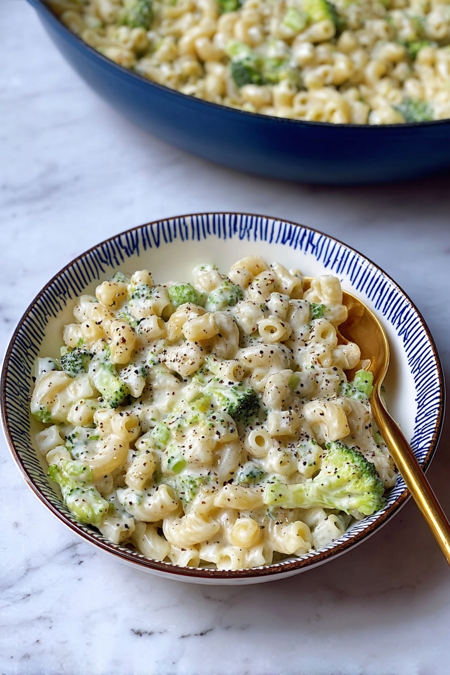 Creamy Broccoli Pasta, creamy broccoli pasta recipe, cheesy broccoli pasta, easy broccoli pasta dinner, quick vegetarian pasta - A white bowl with a blue rim holds a creamy pasta dish made with small tube-shaped pasta mixed with small green broccoli pieces. The pasta is covered in a white, thick sauce, speckled throughout with black pepper. A gold spoon is placed on the right side of the bowl, partially resting in the pasta. Behind the bowl, there is a large blue pan filled with the same creamy pasta and broccoli mixture. The dish is set on a surface with a white marbled texture. photo taken with an iphone --ar 2:3 --v 7