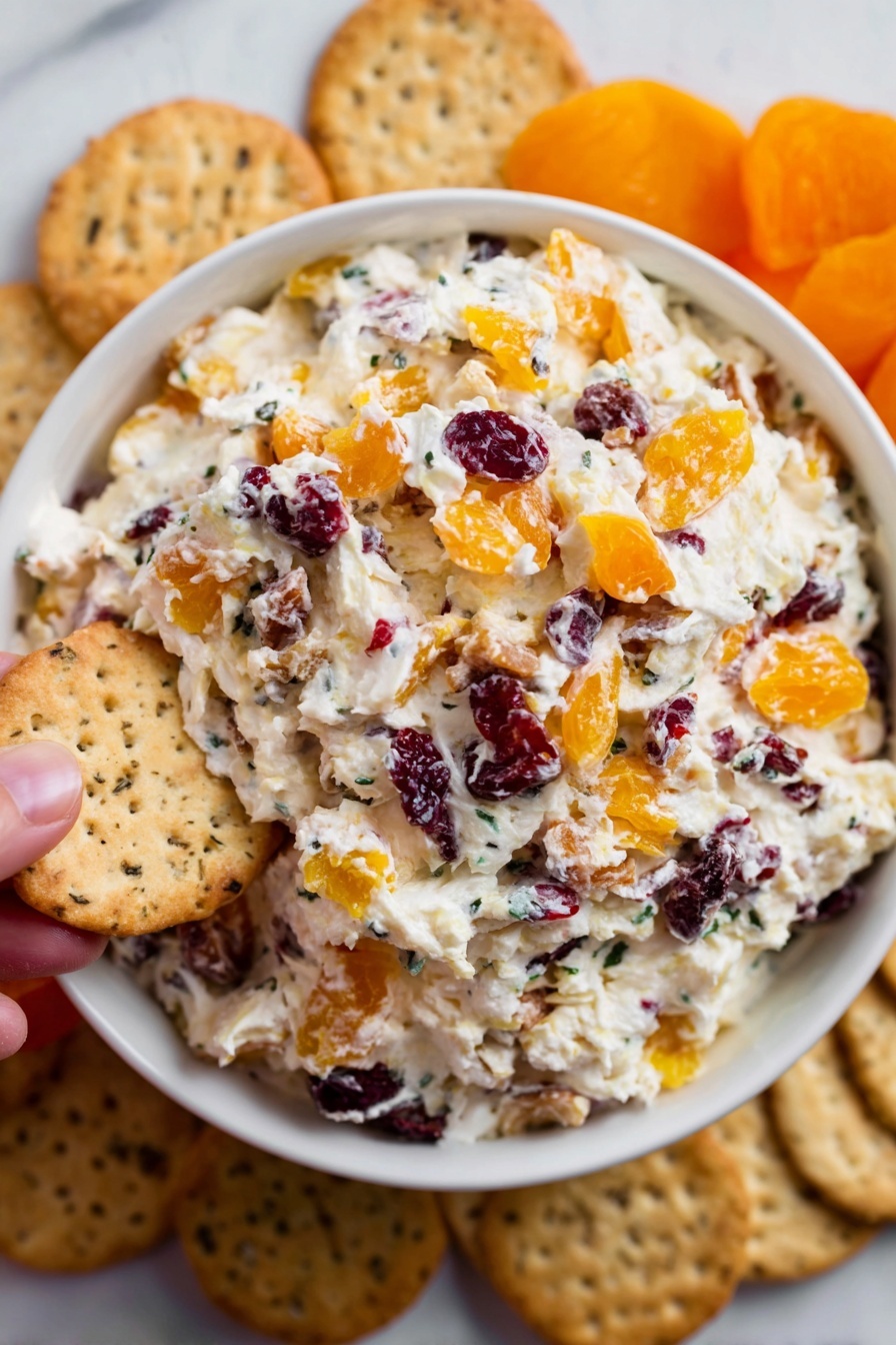 Cranberry Orange Cream Cheese Spread, easy fruit cream cheese spread, holiday appetizer ideas, no-cook fruit spread, flavorful cheese dip - A white bowl is filled with a creamy mixture that has three layers of visible ingredients: the base is soft white cream cheese, mixed throughout with chunks of orange dried apricots and small dark red dried cranberries, creating a colorful and textured look. Around the bowl, there are beige crackers placed on a white marbled surface, some showing a speckled texture. A woman's hand is holding one of the crackers near the edge of the bowl. The scene is bright and clear, showing the fresh, inviting look of the snack. photo taken with an iphone --ar 2:3 --v 7
