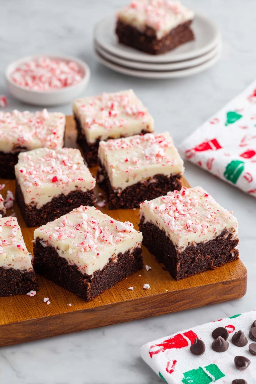 Peppermint Bark Brownies, festive holiday brownies, peppermint chocolate dessert, fudgy brownie recipe, Christmas treat recipes - A wooden board holds eleven square brownies, each with two layers: a dark brown, dense textured base with visible chunks inside, topped by a creamy white layer sprinkled with crushed pink peppermint pieces giving a slightly rough texture. Three brownies are separated at the front, showing clear edges and layers. Behind the board, there is a small white stack of four plates with one brownie on top, and to the right, a white napkin with a pattern of small green and red chef hats rests on a white marbled surface. Scattered peppermint candies and a few chocolate chips decorate the bottom edge of the surface. Photo taken with an iphone --ar 2:3 --v 7