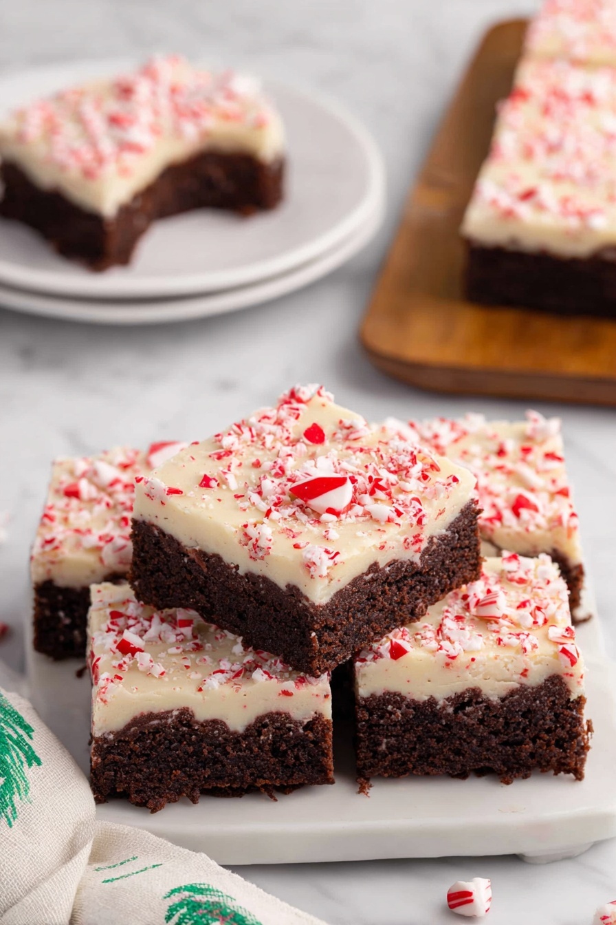 Peppermint Bark Brownies, festive holiday brownies, peppermint chocolate dessert, fudgy brownie recipe, Christmas treat recipes - The image shows a stack of four square peppermint brownies on a white plate with a white marbled surface underneath. Each brownie has two layers: a thick, dark brown chocolate base with a moist, dense texture, and a creamy white topping sprinkled with crushed red peppermint bits that add a pop of color and texture. One brownie on top has a small bite taken out, revealing the rich chocolate layer inside. In the background, there is a similar white plate with another brownie on it, and a soft white cloth with green patterns is visible on the side. photo taken with an iphone --ar 2:3 --v 7