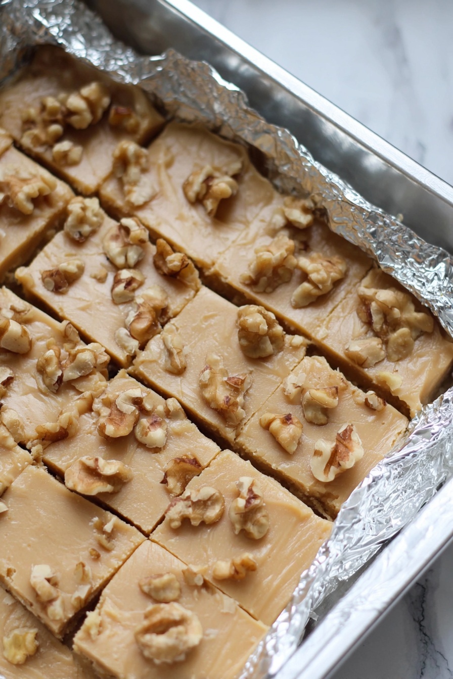 Maple Walnut Fudge, maple walnut fudge recipe, homemade maple fudge, easy fudge with walnuts, cozy maple walnut treats - The image shows a baking pan lined with crinkled aluminum foil holding a light beige fudge cut into small square pieces, topped with scattered walnut chunks. The fudge has a smooth but slightly cracked texture with the nuts unevenly spread on top. The pan rests on a white marbled surface. photo taken with an iphone --ar 2:3 --v 7