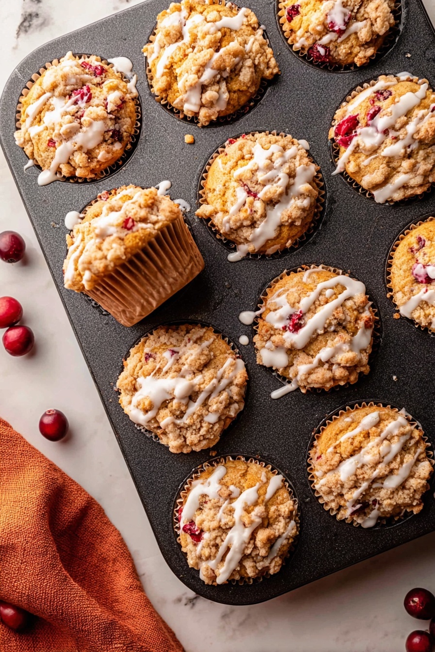 Cranberry Orange Muffins, cranberry orange muffin recipe, citrus breakfast muffins, quick cranberry muffins, easy fall muffin recipe - The image shows a dark muffin tin with nine muffins inside it, all golden brown with a crumbly topping that is light tan and unevenly spread over each muffin. Bright red berry pieces peek through the muffins, adding spots of color. White glaze is drizzled in thin lines over the top of each muffin, contrasting with the crumbly texture. One muffin is pulled slightly out of its spot showing the sides, which have a ridged paper liner of a light brown color. The muffin tin rests on a white marbled surface with a soft orange cloth partially visible in the bottom left corner and a few dark red whole berries scattered near the tin. photo taken with an iphone --ar 2:3 --v 7