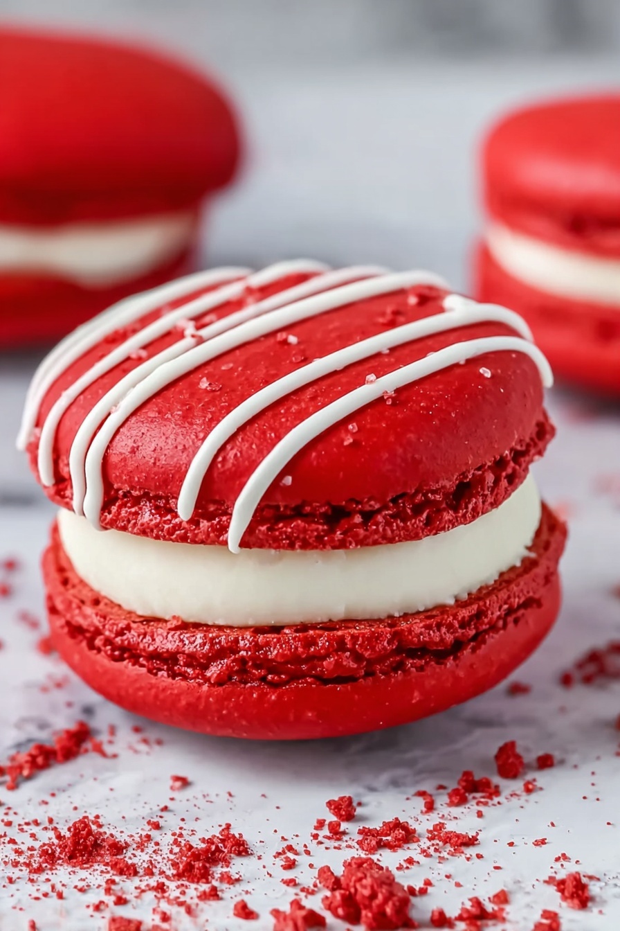Red Velvet Macarons with Cream Cheese Frosting, Red Velvet Macaron Recipe, Homemade Red Velvet Macarons, Easy Macaron Filling, Elegant Dessert for Parties - The image shows a close-up of a bright red macaron with three visible layers: two smooth, round, glossy red shells with a slightly rough edge texture, and a thick white cream filling in the middle. The top shell is decorated with thin white icing lines running diagonally. The macaron rests on a white marbled surface with some white drizzle and red crumbs around it, adding detail and texture. In the background, there is a blurred second red macaron. Photo taken with an iphone --ar 2:3 --v 7