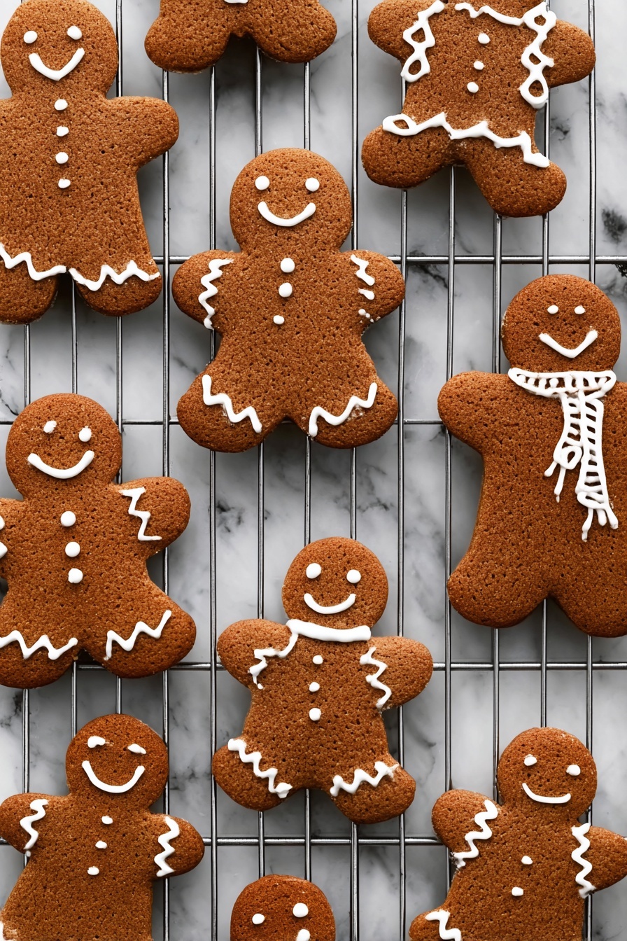 Gingerbread Cookies, gingerbread cookies recipe, holiday cookie ideas, spiced cookies, easy gingerbread treats - The image shows several gingerbread cookies shaped like people, placed on a metal cooling rack over a white marbled texture. Each cookie is a warm brown color with a rough texture and decorated with white icing in different patterns: some have smiles, eyes, buttons, and zigzag designs on arms and legs, while others have scarves or simple outlines on the edges. The cookies are arranged in various sizes and angles across the cooling rack, creating a cozy and festive feel. photo taken with an iphone --ar 2:3 --v 7