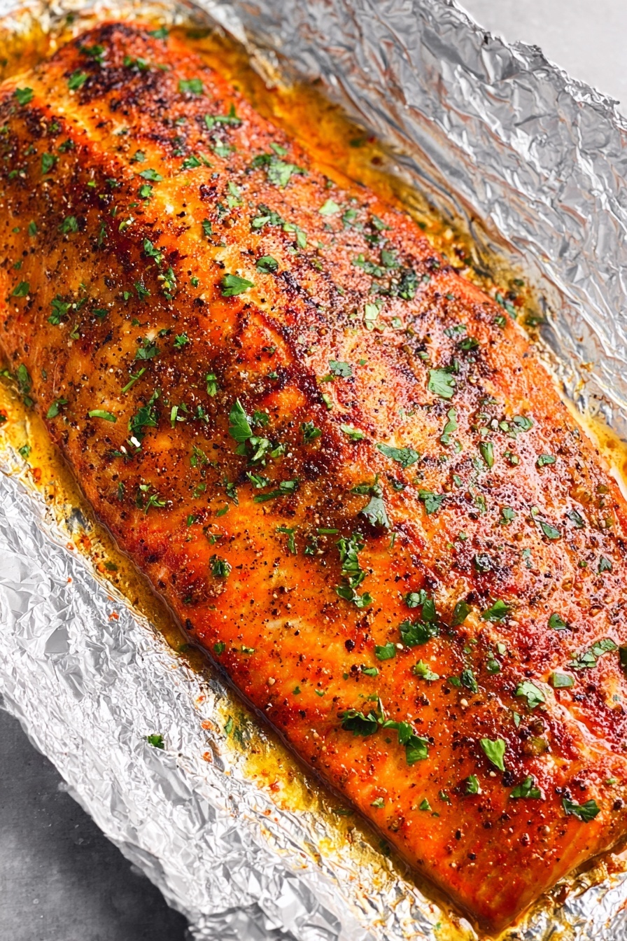 Oven Baked Salmon with Garlic and Herbs, healthy baked salmon, easy salmon dinner, flavorful salmon recipe, quick seafood recipes - A large piece of cooked salmon lies on crinkled aluminum foil in a baking pan, with a golden-brown seasoned crust on top sprinkled with green herbs. The salmon is flaked in the lower right area, showing moist, soft, pink layers inside with some parts breaking apart. A silver fork rests next to the flaked salmon on the right side. The baking pan sits on a white marbled surface. photo taken with an iphone --ar 2:3 --v 7