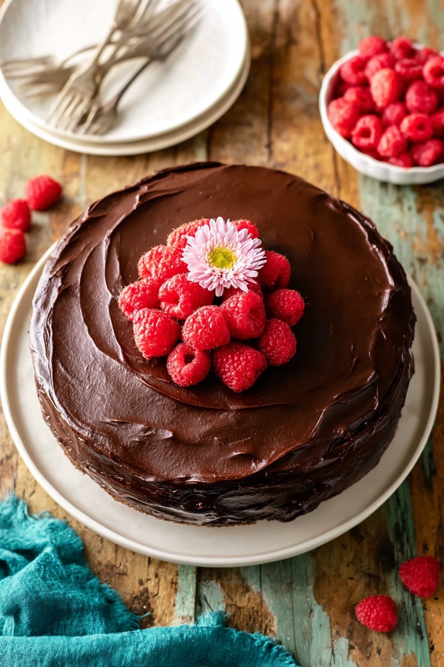 Chocolate Raspberry Cake, chocolate raspberry dessert, easy chocolate cake with raspberry filling, moist chocolate cake recipes, festive berry chocolate cake - A round chocolate cake with two visible layers is covered with thick, smooth dark chocolate frosting. On top, there is a small pile of bright red raspberries with a single light pink flower placed in the center. The cake is sitting on a white plate on a rough wooden surface with a teal cloth partly visible on the bottom left. A small white bowl filled with more raspberries is seen in the top right corner. A white plate with two forks is placed near the top left. Photo taken with an iphone --ar 2:3 --v 7