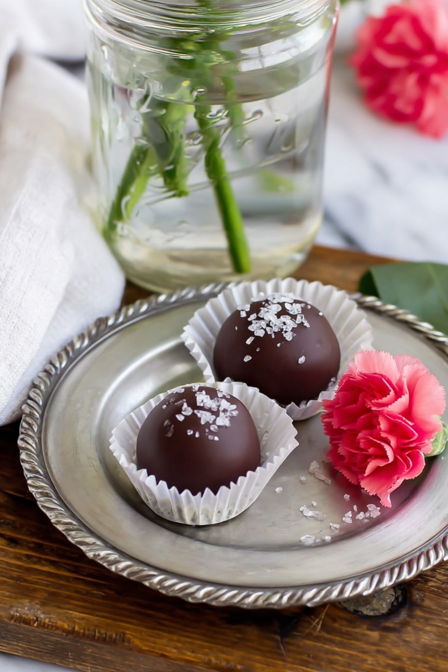 Strawberry Buttercream Candies, strawberry candies recipe, chocolate strawberry treats, fruity chocolate confections, homemade strawberry chocolates - The image shows two round chocolate balls on a small silver plate with scalloped edges. Each chocolate ball is smooth and dark brown, topped with white sugar crystals. One chocolate ball sits in a white paper cup liner, while the other lies directly on the plate. Next to the chocolates is a small bright pink carnation flower with a green stem, slightly touching the plate. The plate is placed on a wooden surface with a white marbled texture background partly visible. A clear glass jar with water and green stems is positioned behind the plate, and part of a white cloth napkin is seen near the jar photo taken with an iphone --ar 2:3 --v 7