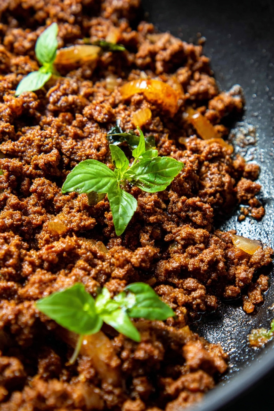 Thai Basil Beef Rolls, Thai Basil Beef Wraps, Thai Beef Lettuce Wraps, Easy Thai Beef Appetizer, Quick Thai Beef Snacks - This close-up image shows a single layer of cooked ground meat, rich brown in color with a slightly crumbly texture, mixed with small pieces of cooked onions that are golden yellow and translucent. Two small green basil sprigs with fresh leaves sit on top of the meat, adding a pop of bright green color. The food is in a black pan, which contrasts with the bright colors of the ingredients. The cooking oil shines slightly on the pan's surface, making the beef look moist and flavorful. Photo taken with an iphone --ar 2:3 --v 7