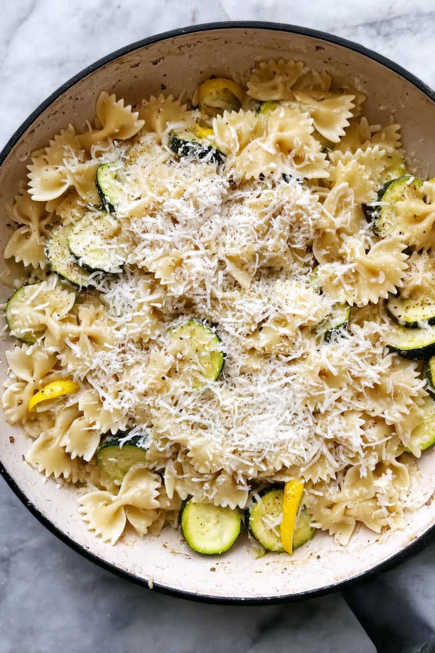 Lemon Chicken Pasta, quick lemon chicken pasta, easy chicken pasta recipes, healthy lemon pasta, simple weeknight pasta dinners - A white pan filled with farfalle pasta mixed with slices of yellow and green zucchini. The pasta and zucchini are covered with a thick layer of finely grated white cheese on top. The mix has a light sauce giving it a slightly shiny look. The pan is placed on a white marbled surface. photo taken with an iphone --ar 2:3 --v 7