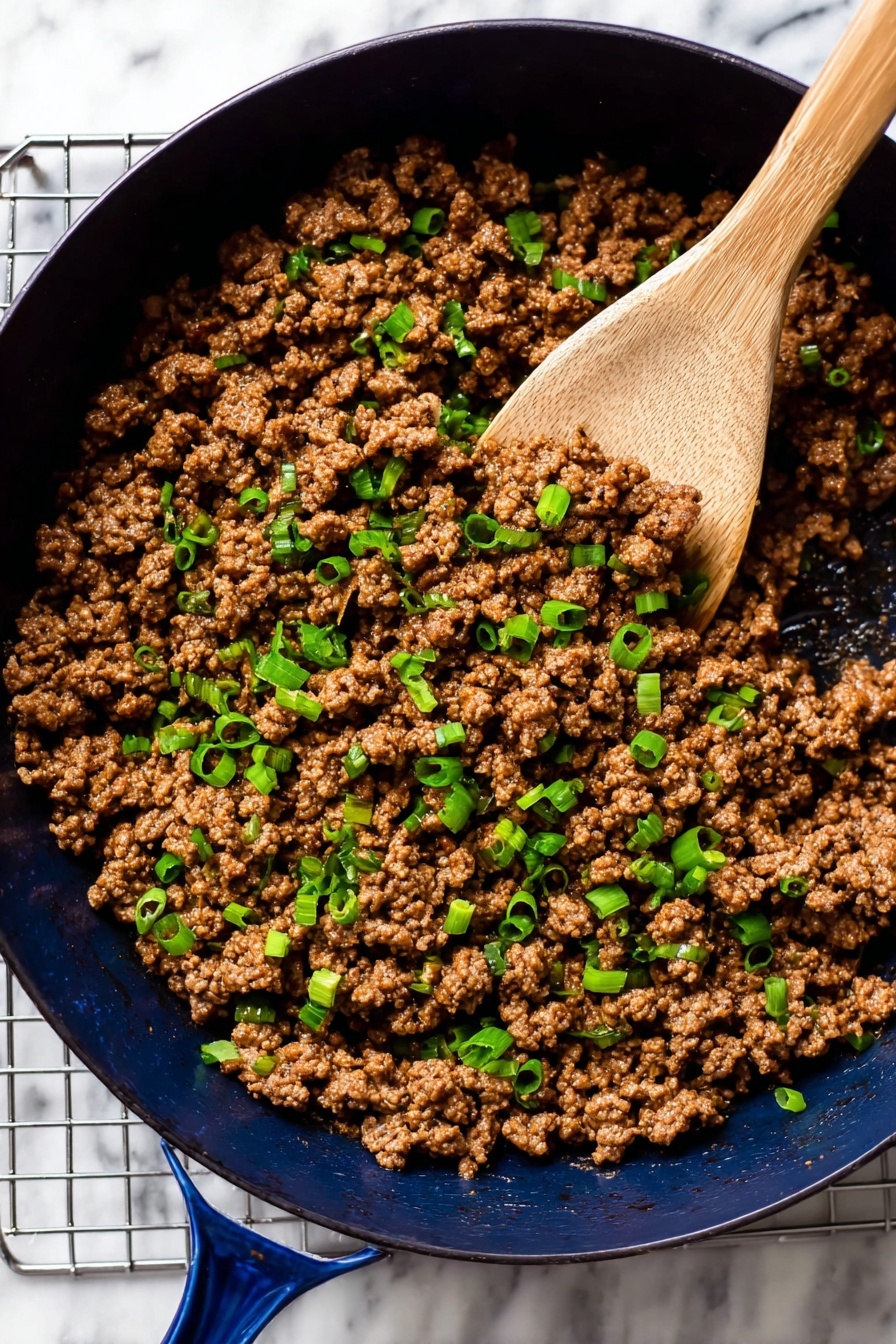 Korean Beef Bowl, Korean Beef Bowl recipe, quick Korean Beef Bowl, easy Korean Beef Bowl, savory Korean beef stir-fry - A dark blue pan filled with cooked ground meat mixed with small pieces of green onion scattered evenly on top. The meat is rich brown with a soft texture, and the green onions add bright green spots throughout. A wooden spoon is scooping some of the meat and onions, positioned on the right side of the pan. The pan sits on a grid metal rack over a white marbled surface. Photo taken with an iphone --ar 2:3 --v 7