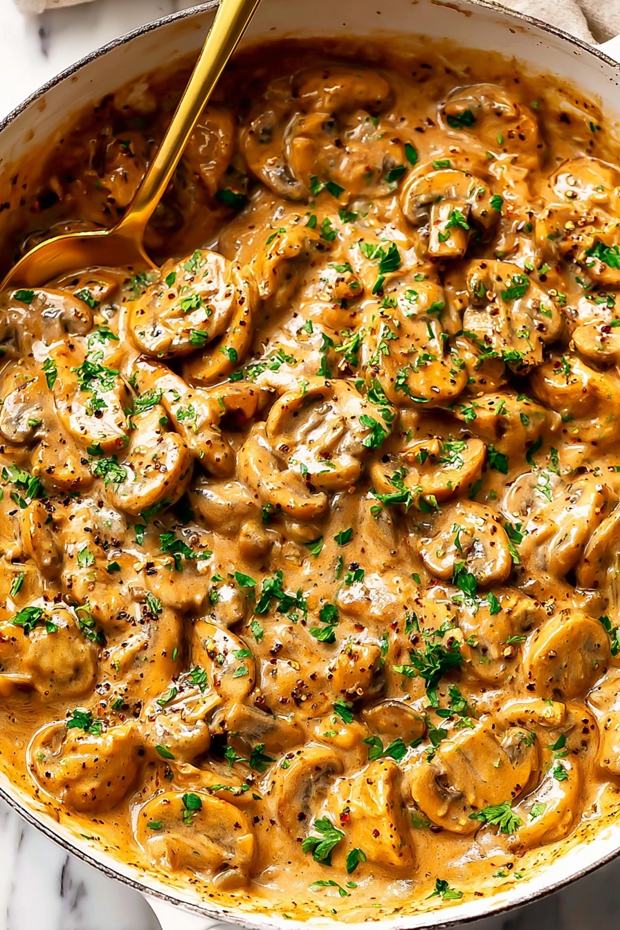 French Onion Pasta with Melted Gruyere, French Onion Pasta, Gruyere Pasta Recipe, Caramelized Onion Pasta, Comforting Pasta Dishes - The image shows a large white pan filled with a creamy mushroom dish. The sauce is thick and light brown with visible specks of black pepper and herbs. The dish contains sliced mushrooms that are golden with a soft texture, mixed evenly in the sauce. Fresh green parsley pieces are scattered across the surface, adding contrast to the warm colors. A gold-colored spoon is partially dipped into the food, with its handle resting on the edge of the pan. The background has a white marbled surface. Photo taken with an iphone --ar 2:3 --v 7