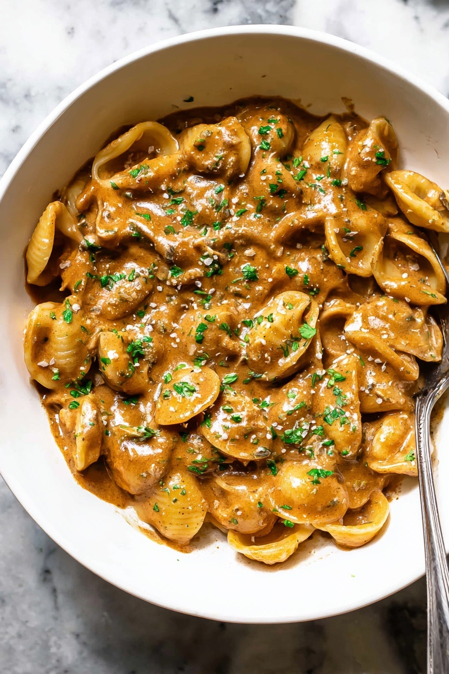 French Onion Pasta with Melted Gruyere, French Onion Pasta, Gruyere Pasta Recipe, Caramelized Onion Pasta, Comforting Pasta Dishes - A white bowl filled with about three layers of small, round pasta shells covered in a thick, creamy, brown sauce with a smooth texture. The sauce coats each piece of pasta evenly, giving a shiny look. Chopped green herbs are sprinkled all over the top layer, adding small spots of bright green contrast. There is a silver spoon partly dipped into the pasta on the right side of the bowl. The bowl sits on a white marbled surface. Photo taken with an iphone --ar 2:3 --v 7