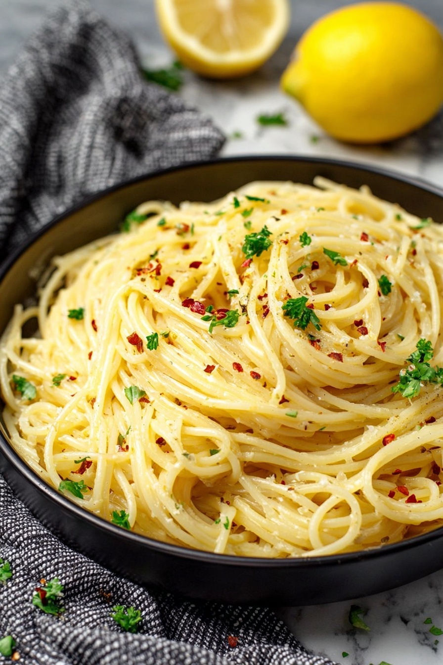 Lemon Garlic Pasta Sauce, lemon garlic pasta sauce recipe, easy lemon garlic pasta, quick pasta sauce with lemon and garlic, fresh pasta sauce for dinner - A close-up view of a single layer of creamy spaghetti pasta in a round black bowl, with the noodles showing a smooth and slightly shiny texture. The spaghetti is sprinkled with small bits of green parsley and red chili flakes that add color contrast. In the background, there is a whole lemon and a half lemon placed slightly out of focus. The bowl sits on a white marbled surface with a gray and white checkered cloth draped casually beside it. The overall look is fresh and inviting, with soft natural lighting. photo taken with an iphone --ar 2:3 --v 7